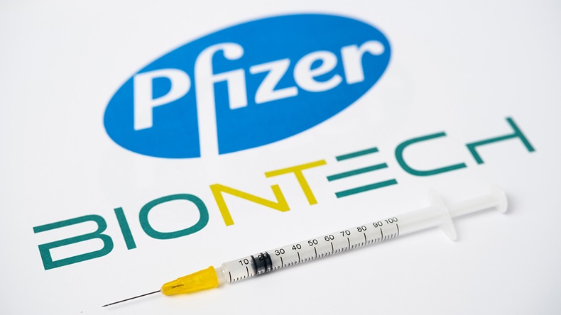 Stafford / United Kingdom - November 9 2020: Pfizer BioNTech Covid-19 vaccine concept. Syringe balanced on a fingertip and blurred company logos on the background. Selective focus.