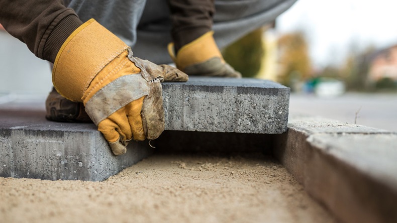 Workman laying exterior paving stones in a low angle view of his gloved hands fitting a brick into a tight fitting space on a sand foundation.