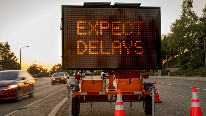 Mobile Electronic Traffic Sign stating “expect Delays” taken at sunset with traffic blurred driving past the sign and traffic cones