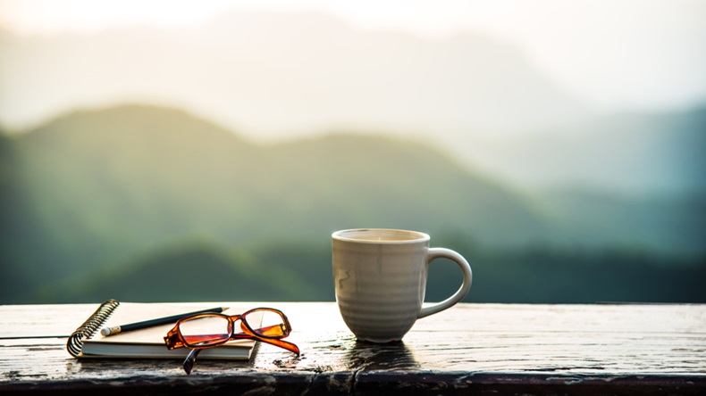 A coffee cup, eyeglasses and a note book on wooden table with sunrise and mountain bokeh background