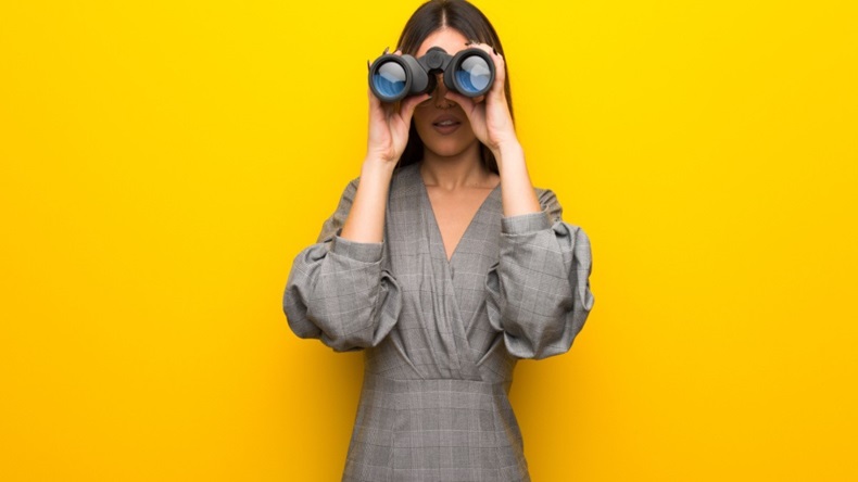 Businesswoman looking through a pair on binoculars against a yellow background