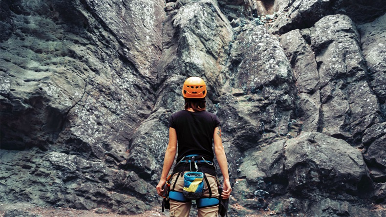 Young woman wearing in climbing equipment standing in front of a stone rock outdoor and preparing to climb, rear view
